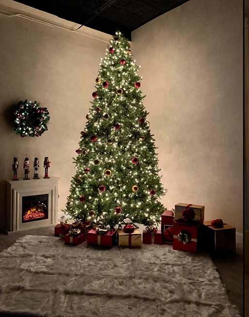 Warm rustic Christmas set at La Vie Rental Studio Clearwater FL — traditional green tree with red ornaments, white electric fireplace, wooden nutcrackers, wreath and wrapped presents on a plush white rug